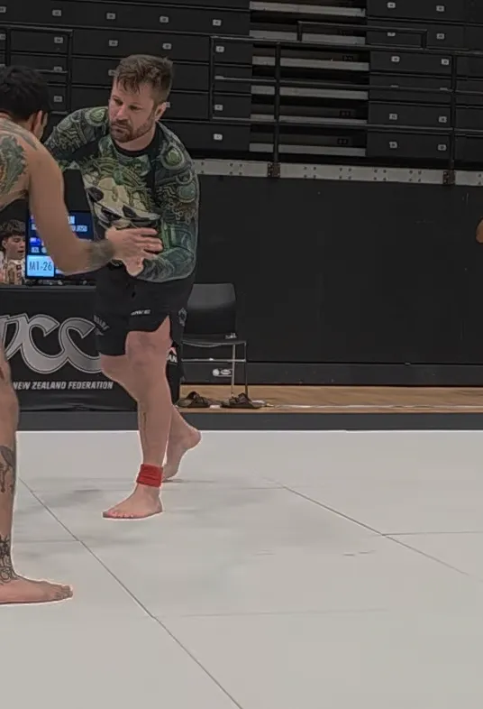 Nick in a patterned rash guard and black shorts, gripping an opponent's wrist on the mat at a New Zealand grappling competition