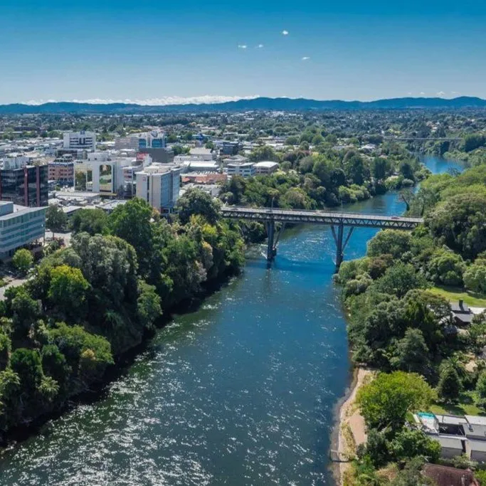 An aerial view of Hamilton, New Zealand, looking along the Waikato River as it runs past the city centre, with a steel arch bridge crossing it and hills on the horizon